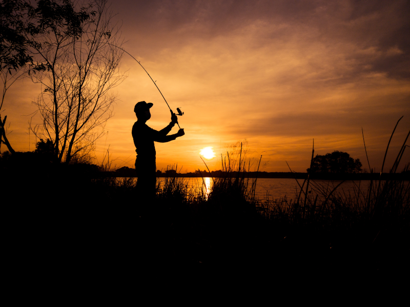 A silhouette of a man on a river bank holding a fishing rod during sunset